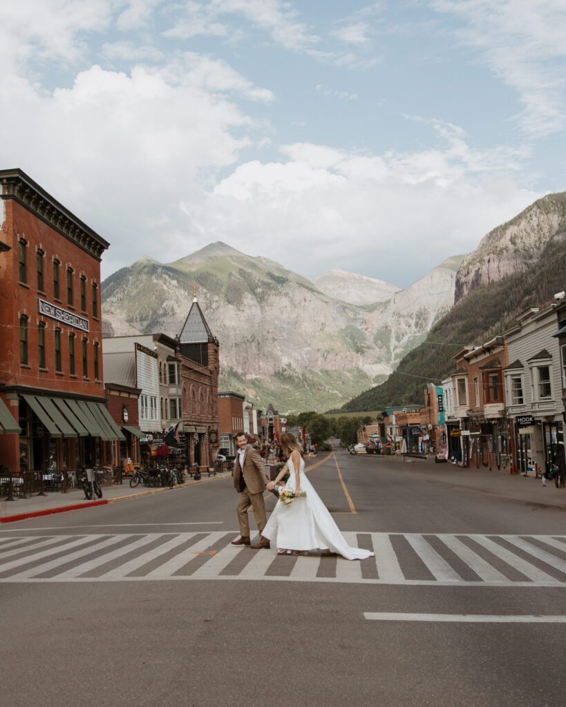 Couple holding hands and walking across a crosswalk in downtown Telluride, surrounded by colorful historic buildings and mountain peaks.